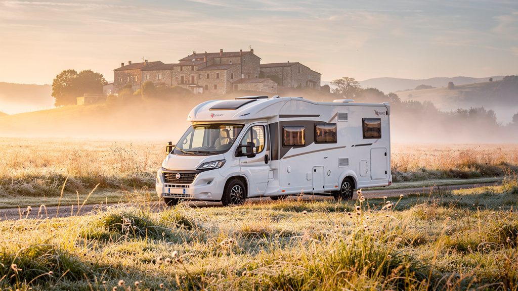 Camping-car familial blanc stationné dans une aire rurale au lever du soleil, entouré de prairies, avec un village en pierre au loin.