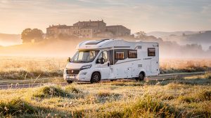 Camping-car familial blanc stationné dans une aire rurale au lever du soleil, entouré de prairies, avec un village en pierre au loin.